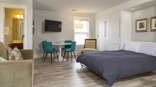 An image of a living room with a Murphy Bed, the bed is currently folded down and on the right side of the room, with a chair to the left side and a dining area at the back of the room