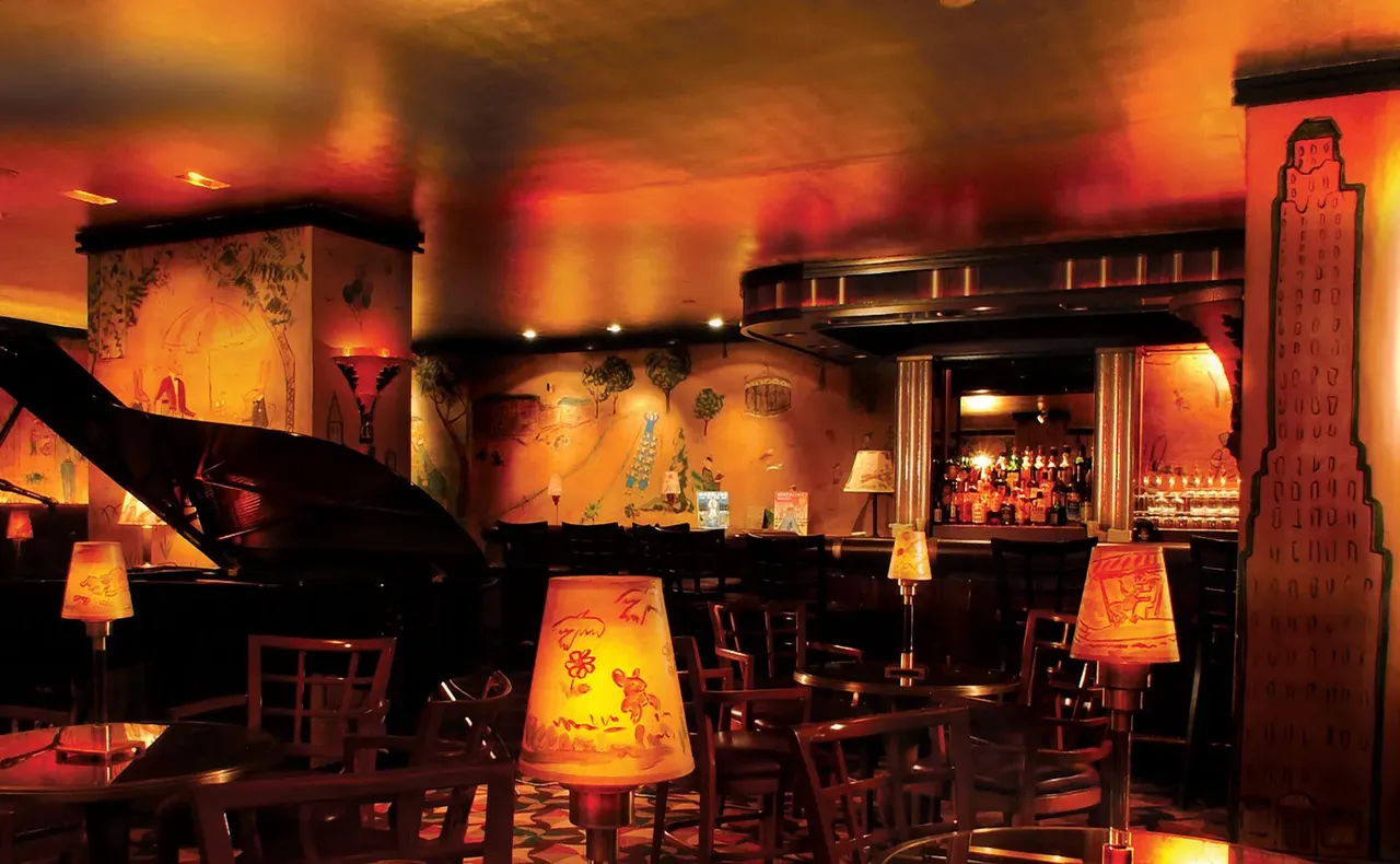 A dimly lit, vibrantly decorated cocktail bar features mural-filled walls and amusing table lamps covered in equally striking drawings.