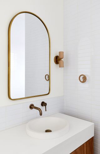 White bathroom with white tiled walls, white countertop, brass tap and mirror and ceramic wall light and towel hook