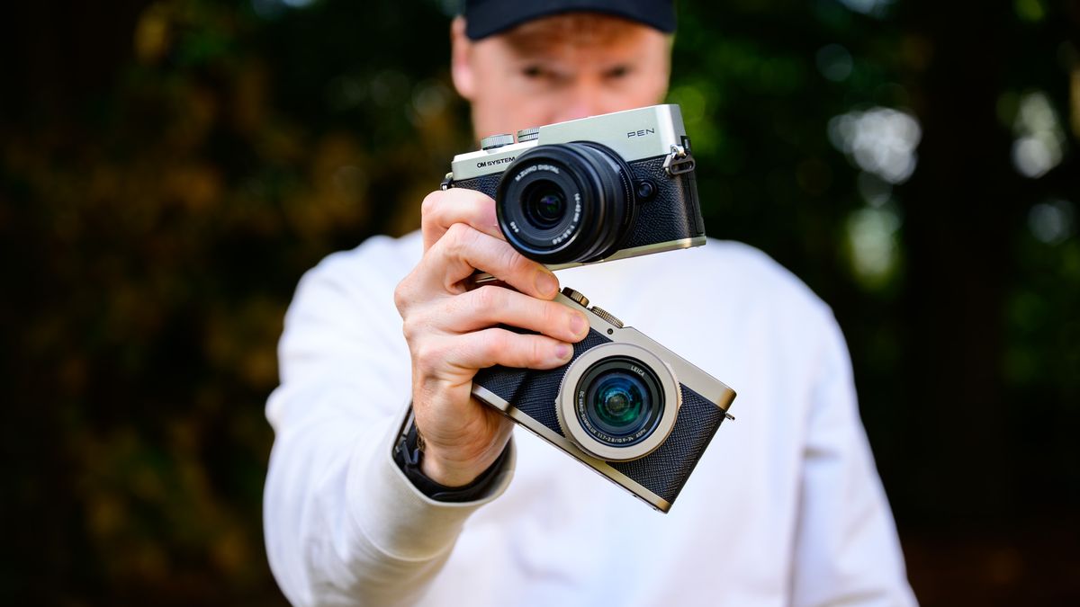 OM System PEN vs Panasonic Lumix L10: two micro four thirds cameras side by side in tech journalist Tim Coleman's hands, dappled tree light background
