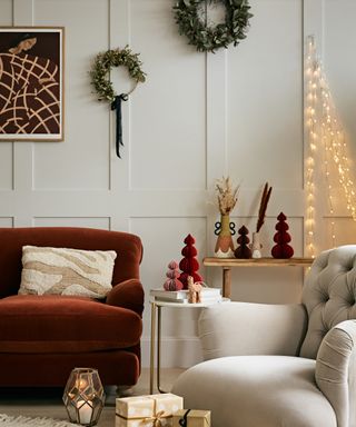 a stunning festive living area, with a red sofa, beige armchair, beige paneled walls, hanging wreaths and festive fairy lights, and presents on the floor