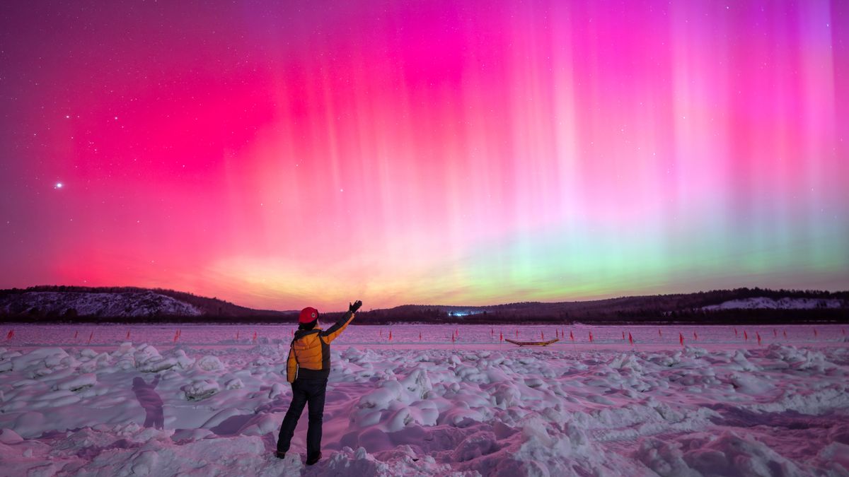 northern lights fill the sky with rich magenta colors and bright pillars of light. a person stands in the foreground of the image and reaches up to the sky with one hand.