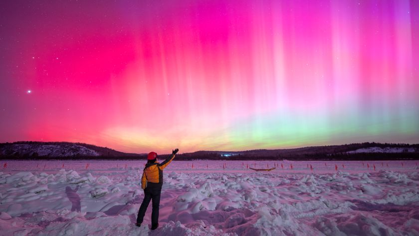 northern lights fill the sky with rich magenta colors and bright pillars of light. a person stands in the foreground of the image and reaches up to the sky with one hand. 