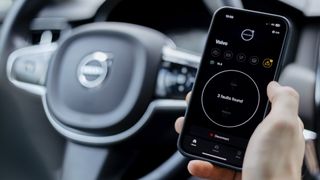 An outstretched hand holding a smartphone with the OBDeleven app on-screen in front of the steering wheel of a car