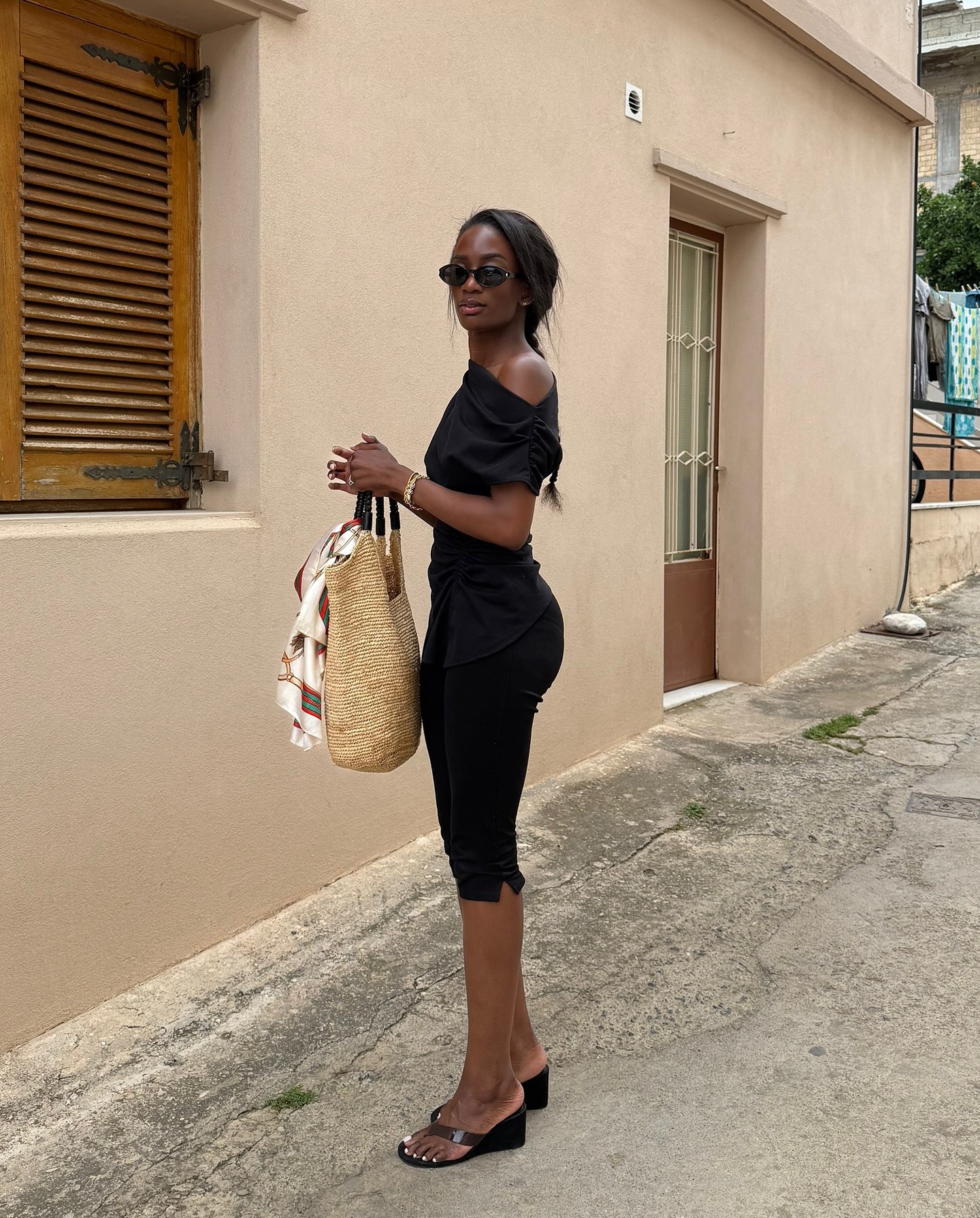 Fashion person Liv Madeline wears a black of the shoulder top, black carpis and black thong sandals and is carrying a crochet tote bag. She is stood in the middle of an old street on a trip away in a recent image from her Instagram.