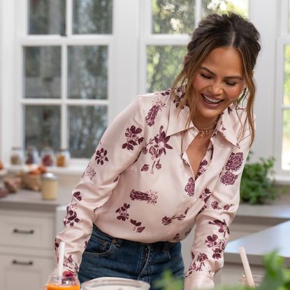 Chrissy Teigen wearing a pink shirt laughing in a white kitchen