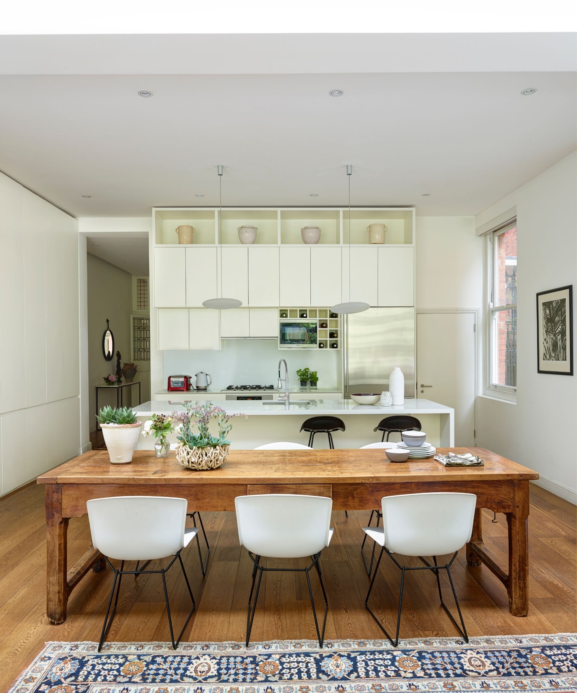 A modern white kitchen space with a large wooden dining table with matching white chairs with black legs, a large patterned rug, a white kitchen island, and white cabinetry.