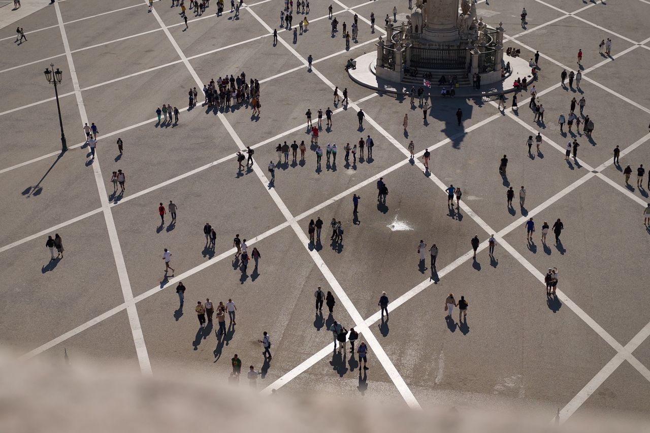 High-angle view of people walking across Pra&ccedil;a do Com&eacute;rcio, casting long shadows across the geometric paving.