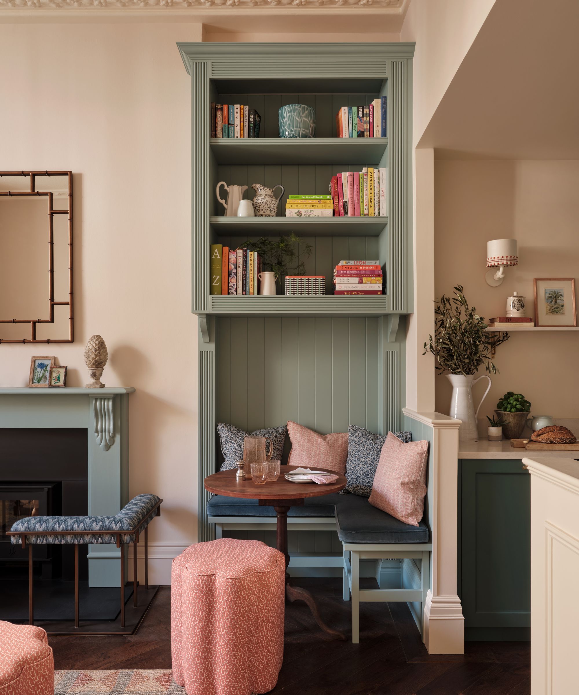 Living room nook with blue painted bookcase, built-in banquette bench with pale blue and pink cushions and wooden round table beside a blue fireplace with fireplace seat and mirror