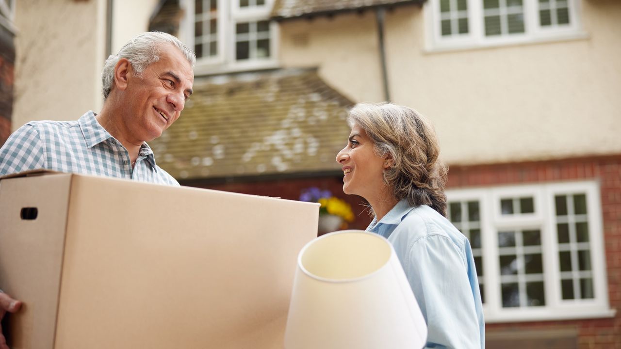 An older couple smile at each other as he holds a moving box and she holds a lamp.