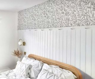 A traditional-style bedroom with tall white wall panelling, by EASipanel, and leaf-themed wallpaper above. There is a wicker-style double bed against the wall, dressed with white bedding. There is also a wall-mounted brass and white bedside light and vase of flowers next to the bed.