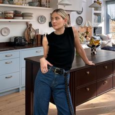 British style influencer Lucy Williams poses in her chic London kitchen with light blue cabinets wearing a simple black sleeveless tank top, woven black belt, and high-waisted jeans