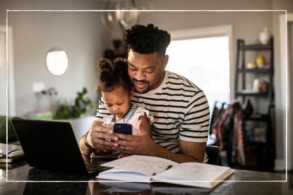 dad sitting at table in front of a laptop looking at his phone, while also holding in toddler daughter in his lap 