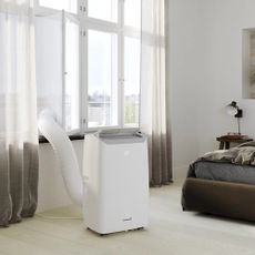 A portable white air conditioner unit in a white living room with a hose disappearing out of the window