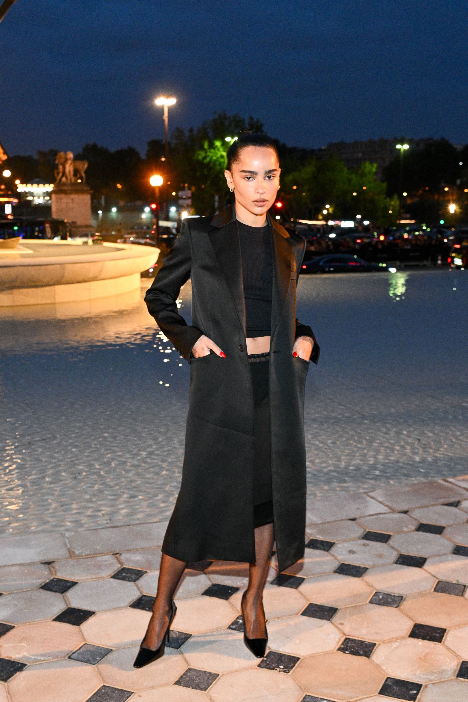 Zoe Kravitz wears a silk long coat, black top, black pencil skirt, and black pumps.