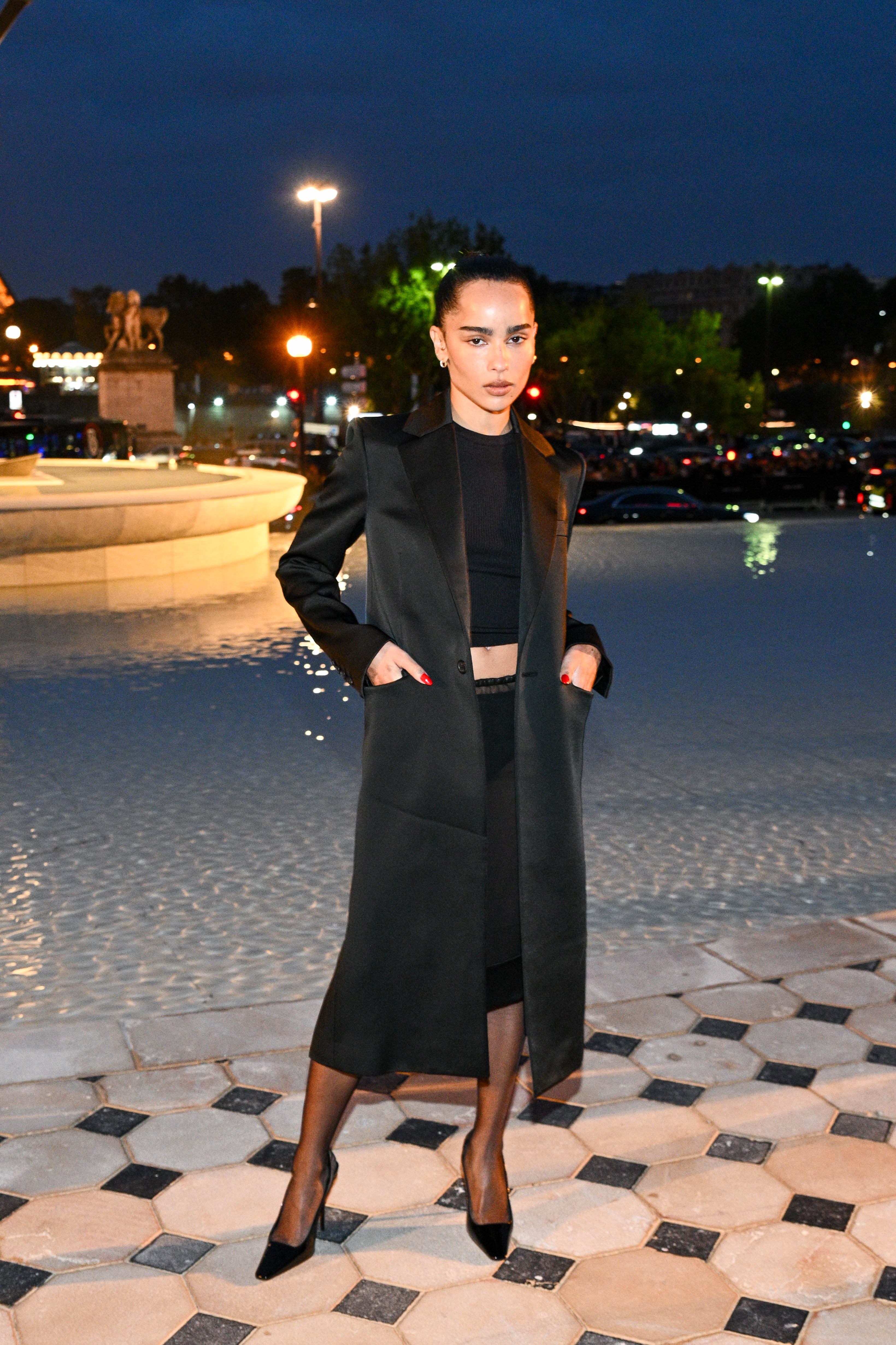 Zoe Kravitz wears a silk long coat, black top, black pencil skirt, and black pumps.