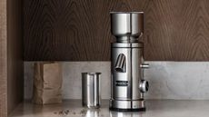 A stainless steel coffee grinder on a grinder on a marble counter with a wooden backsplash.