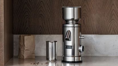 A stainless steel coffee grinder on a grinder on a marble counter with a wooden backsplash.
