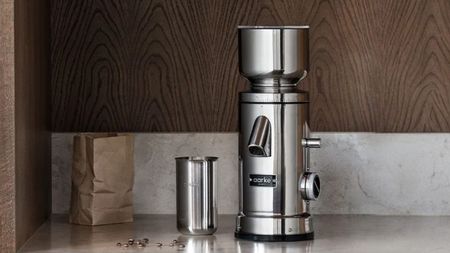 A stainless steel coffee grinder on a grinder on a marble counter with a wooden backsplash.