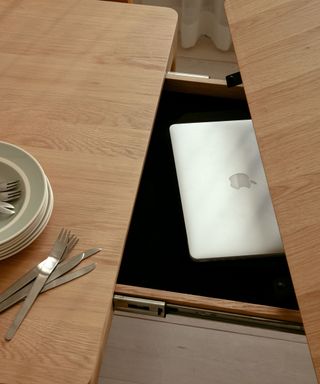 A birds eye view of a wooden dining table with a green plate and a knife and fork set on it, with an open storage drawer with a silver laptop in it