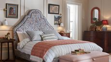 A floral bed frame in a layered, classic bedroom with paneled cream walls, mid century wooden mismatched nightstands and drawer units, with striped bedding, a collection of throw pillows and blankets. There is a pink velvet ottoman at the foot of the bed, and various frames art on the walls