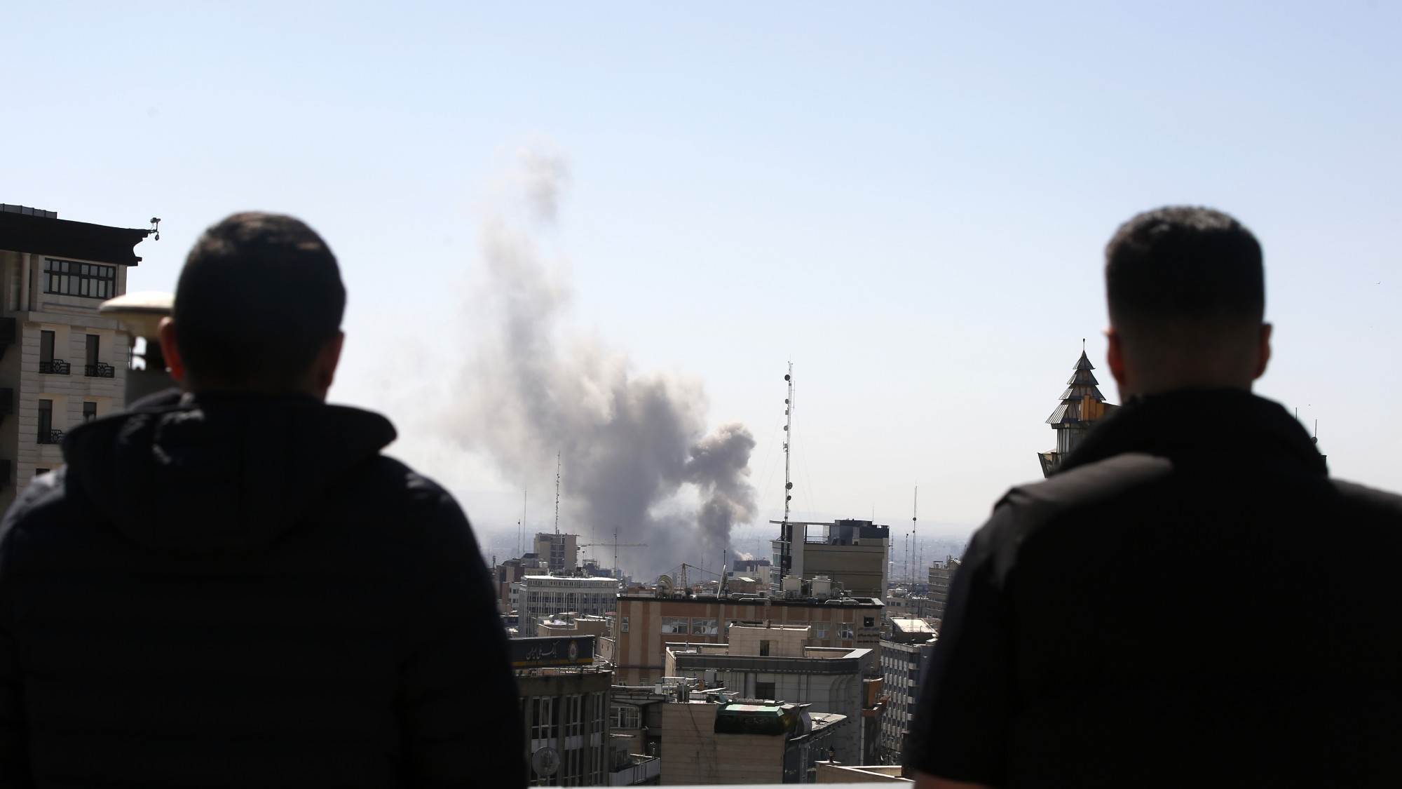Two men watch plumes of smoke rise over a residential area of Tehran