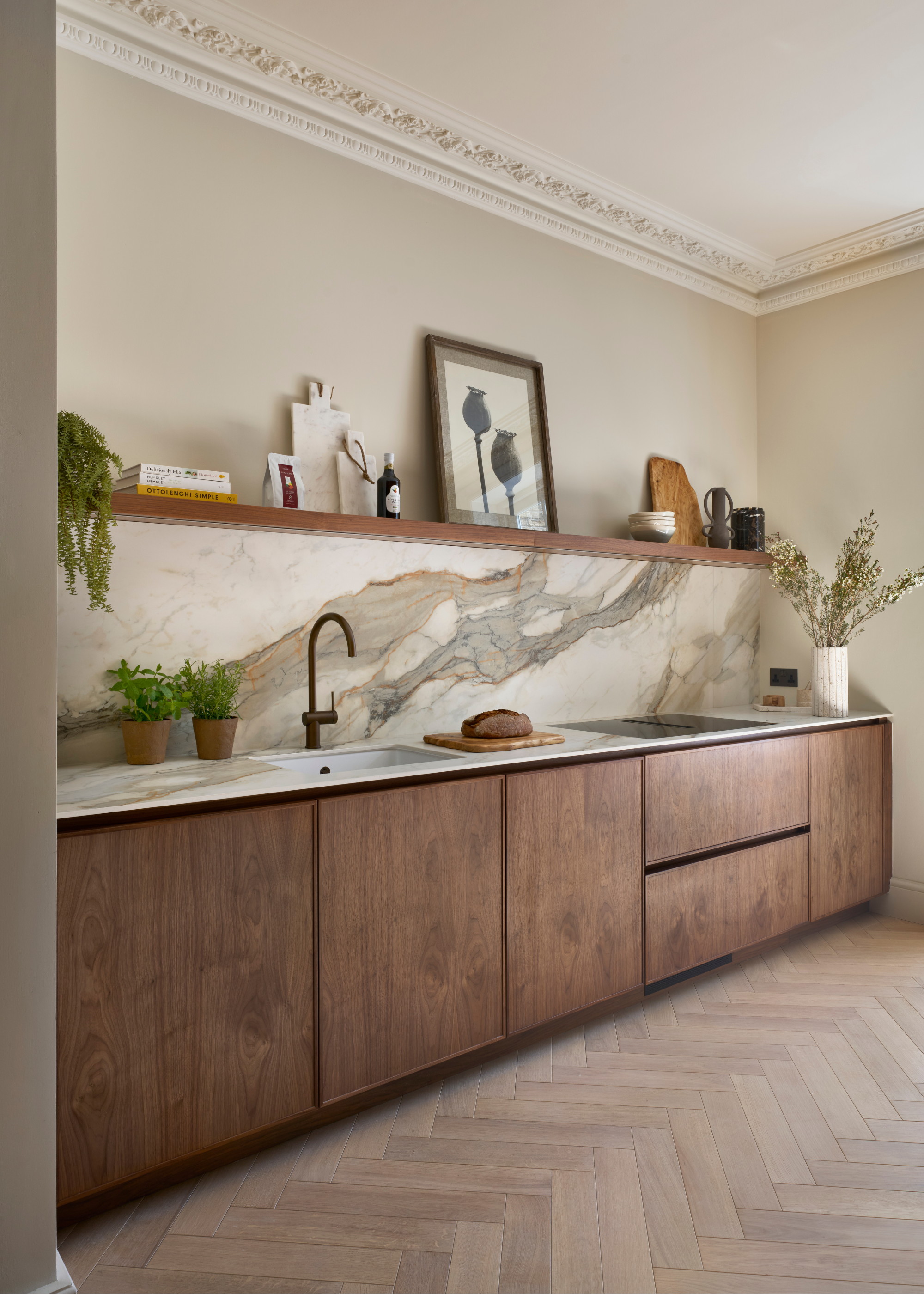 An angled view of an all-wood galley kitchen with a large slab of dramatically veined light marble as the backsplash and open shelving above that with artwork, greenery and other objects