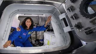 a woman astronaut in a flight suit posing in the doorway of a spacecraft simulator. the open door is at right