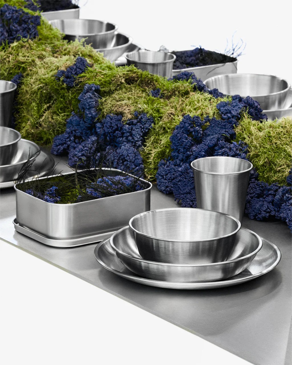 Image of stainless steel serve ware on a stainless steel table with a centerpiece of green and purple flowers.