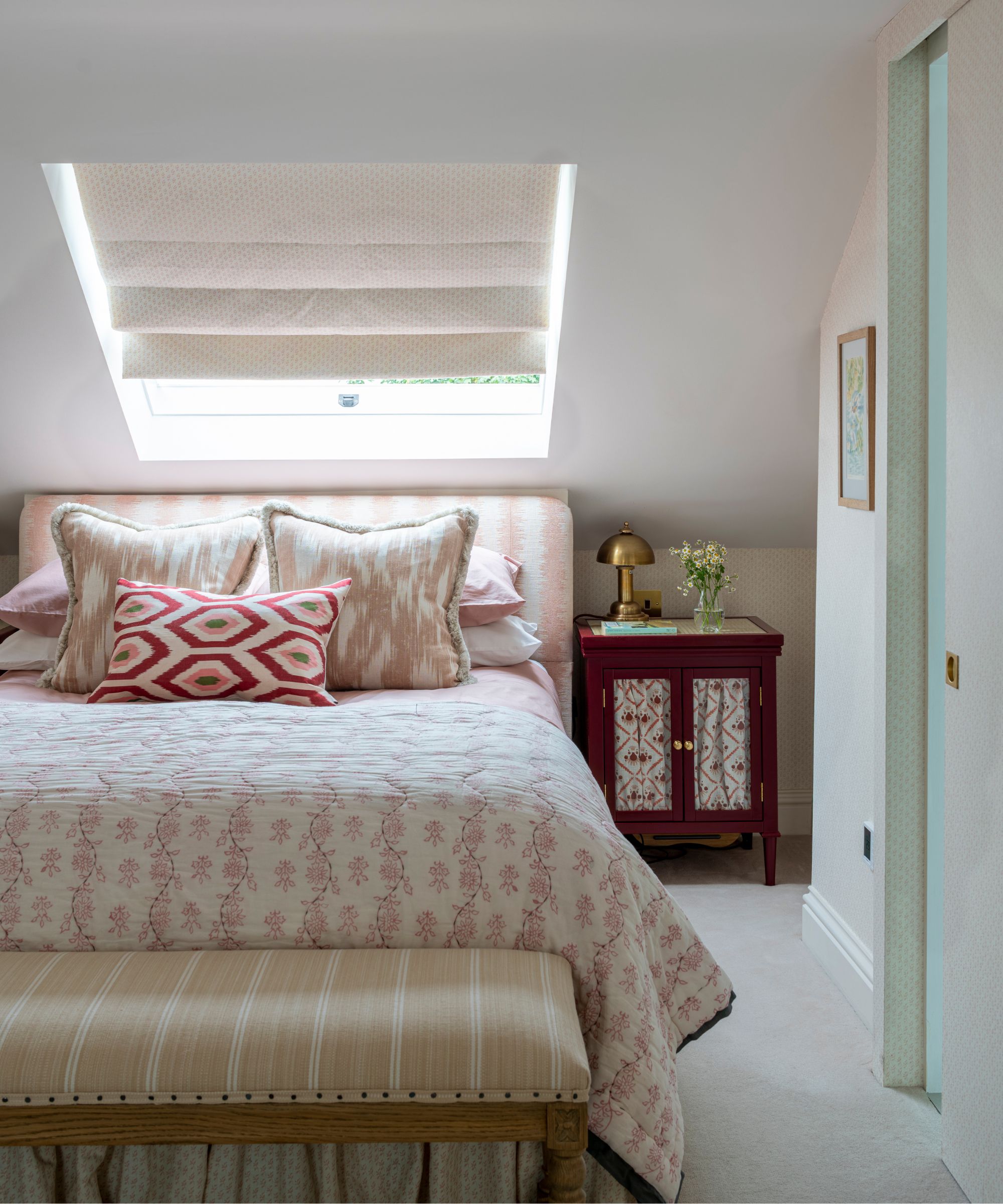 A white attic bedroom with a large double bed with pink and white patterned bedding, cushions and headboard, a striped ottoman, brown bedside table with gold lamp and white blind on a sloped window above the bed.