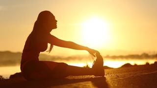 A woman stretching to touch her toes while sitting in front of a rising sun