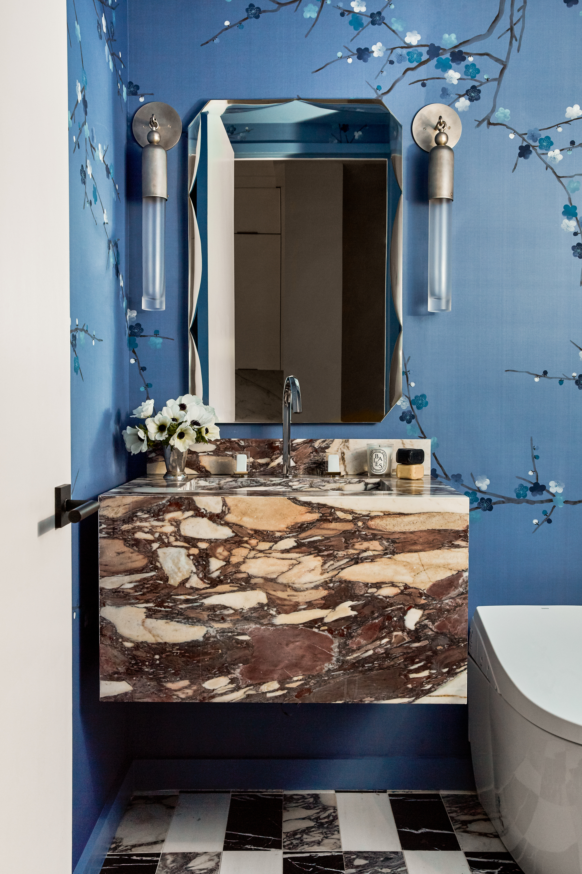 colorful bathroom with blue patterned wallpaper, a brown marble floating vanity, checkered marble floor and wall sconces on either side of mounted wall mirror