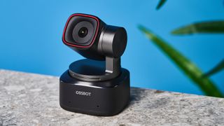 a small PTZ webcam with 4K resolution and a magnetic attachment clip is photographed in front of a blue background and a green plant