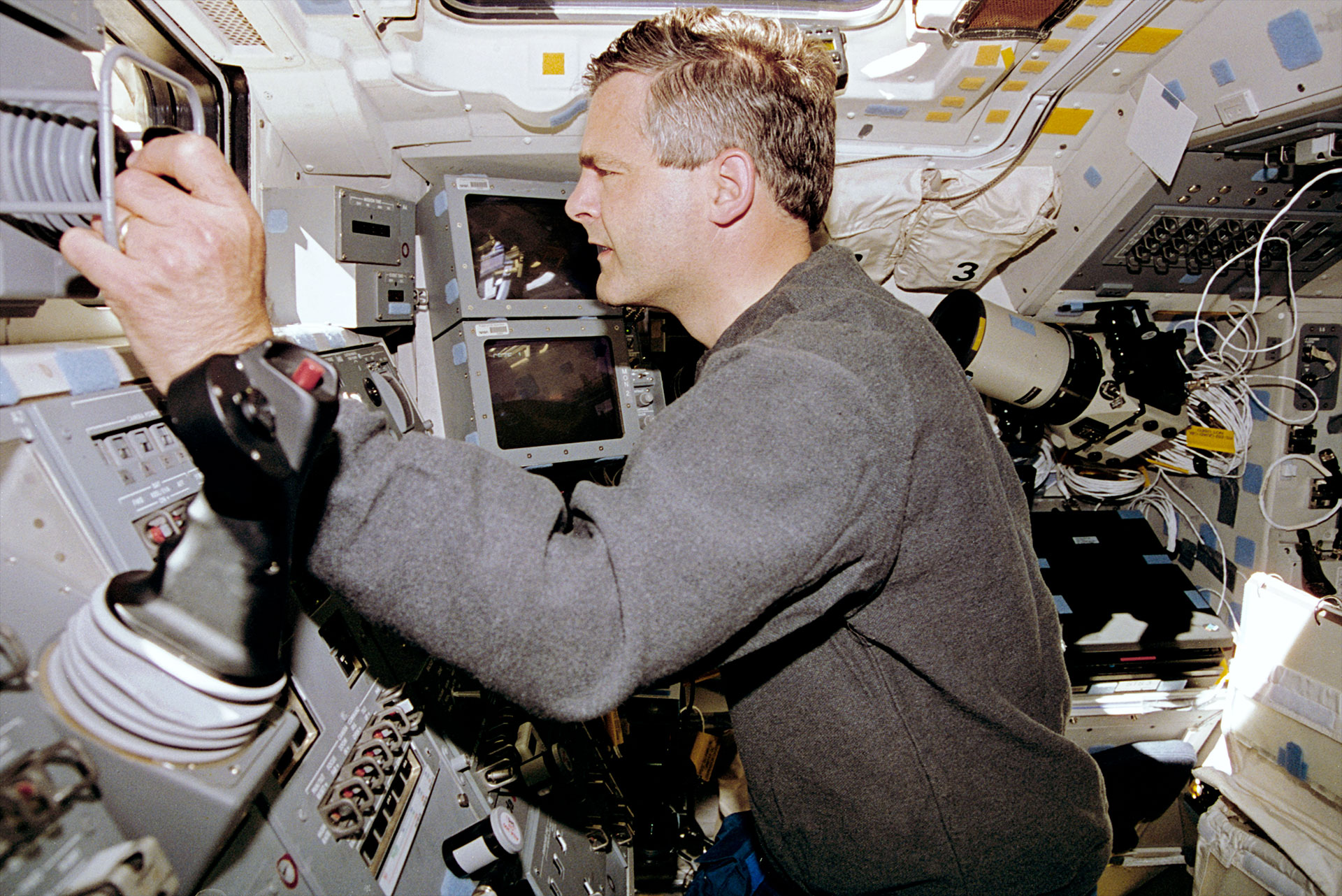A man in a gray sweater with salt and pepper hair works at the controls for a spacecraft