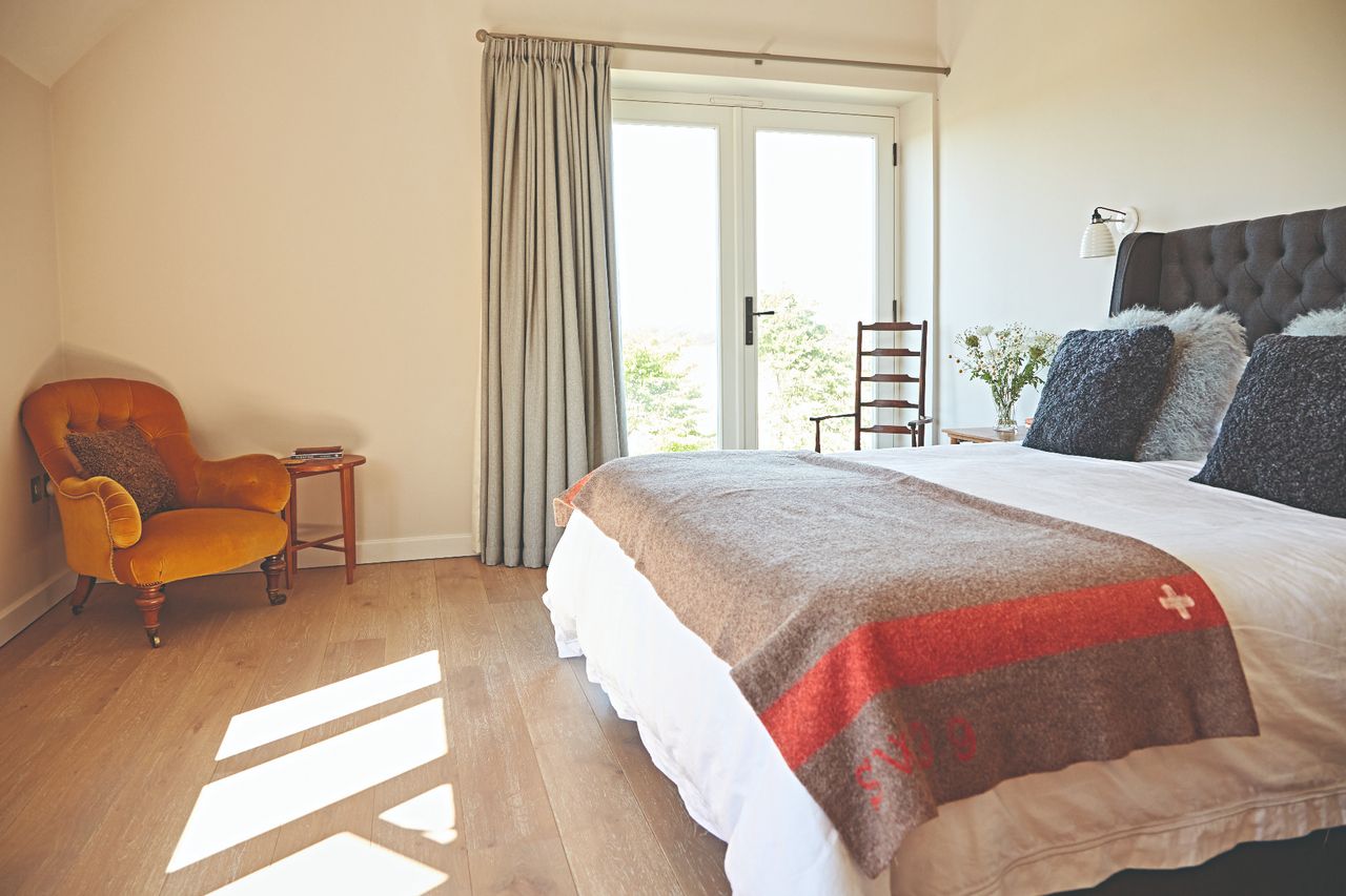 A bedroom with beige walls, french doors, a double bed with cushions and a velvet armchair in the corner