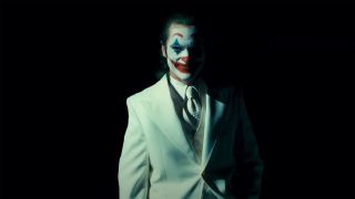 Arthur Fleck smiles with his clown makeup on as he wears a white suit in Joker: Folie a Deux