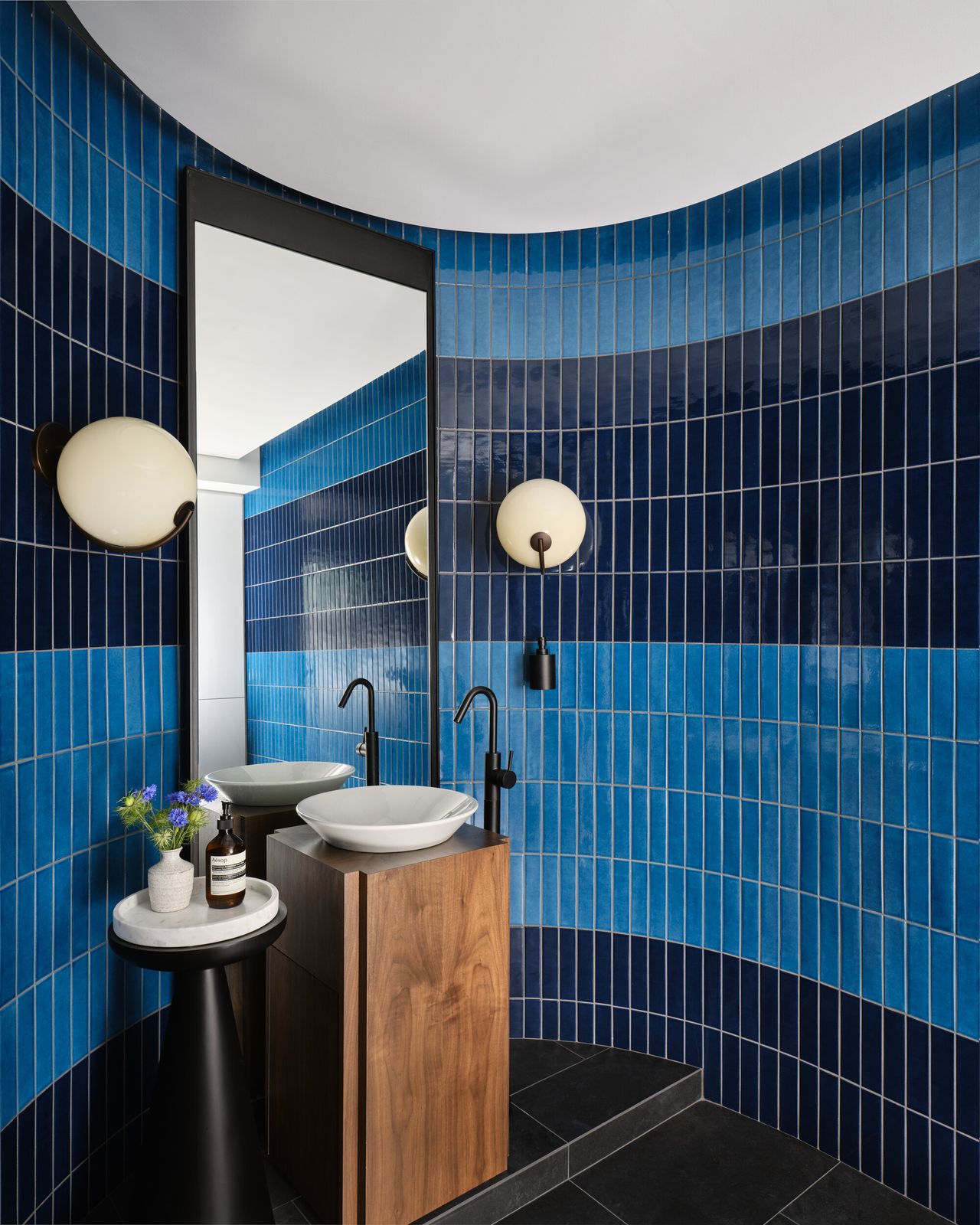 curved bathroom with two tones of blue tiles, a freestanding timber pedestal sink, round wall sconces, a side table with a plate holding hand soap and a vase of flowers, and a wall mirror