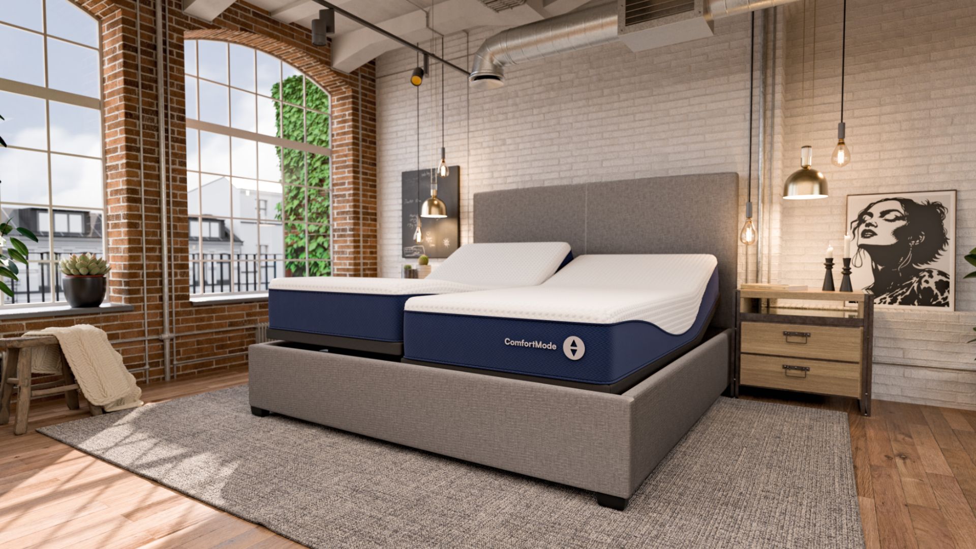 A dual-sided Sleep Number ComfortMode smart bed is shown in a modern, industrial-style bedroom with exposed brick walls and large windows. The bed features two independent mattresses on an adjustable base, with both the head and foot sections slightly elevated. The mattresses have a navy blue base with "ComfortMode" branding and a white, textured top cover. The bed is framed by a grey upholstered headboard and base, set on a grey area rug next to a wooden nightstand with minimalist decor.