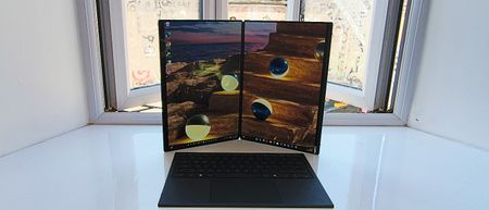 The ASUS Zenbook Duo standing vertically like an open book, displaying a continuous ocean sunset wallpaper split across two portrait screens, with the detached wireless keyboard resting in front.
