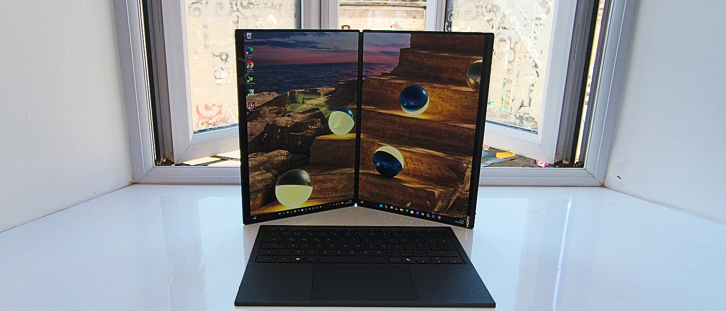 The ASUS Zenbook Duo standing vertically like an open book, displaying a continuous ocean sunset wallpaper split across two portrait screens, with the detached wireless keyboard resting in front.