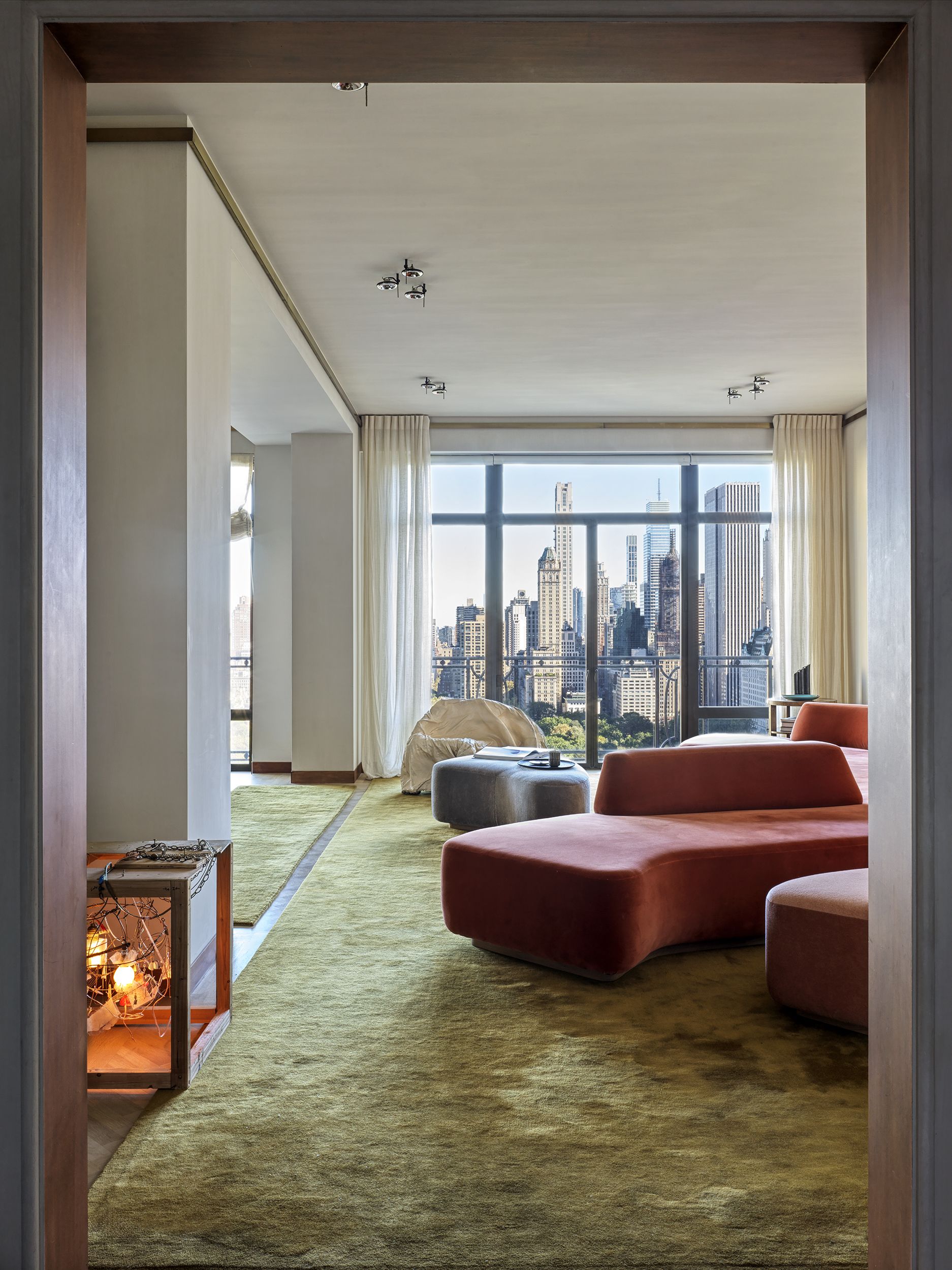 A modern living room with a sweeping view of Central Park with a sculptural red sofa and green carpet, a piece of art on the wall and wooden side tables