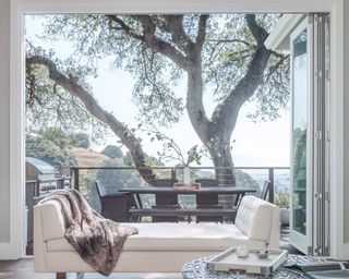 An example of patio door ideas showing a white day bed in front of open bifold doors leading to a terrace and a large tree