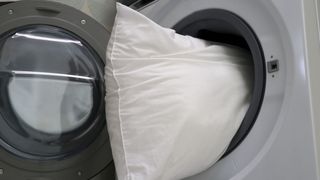 Image shows a white pillow being placed in a washing machine for washing