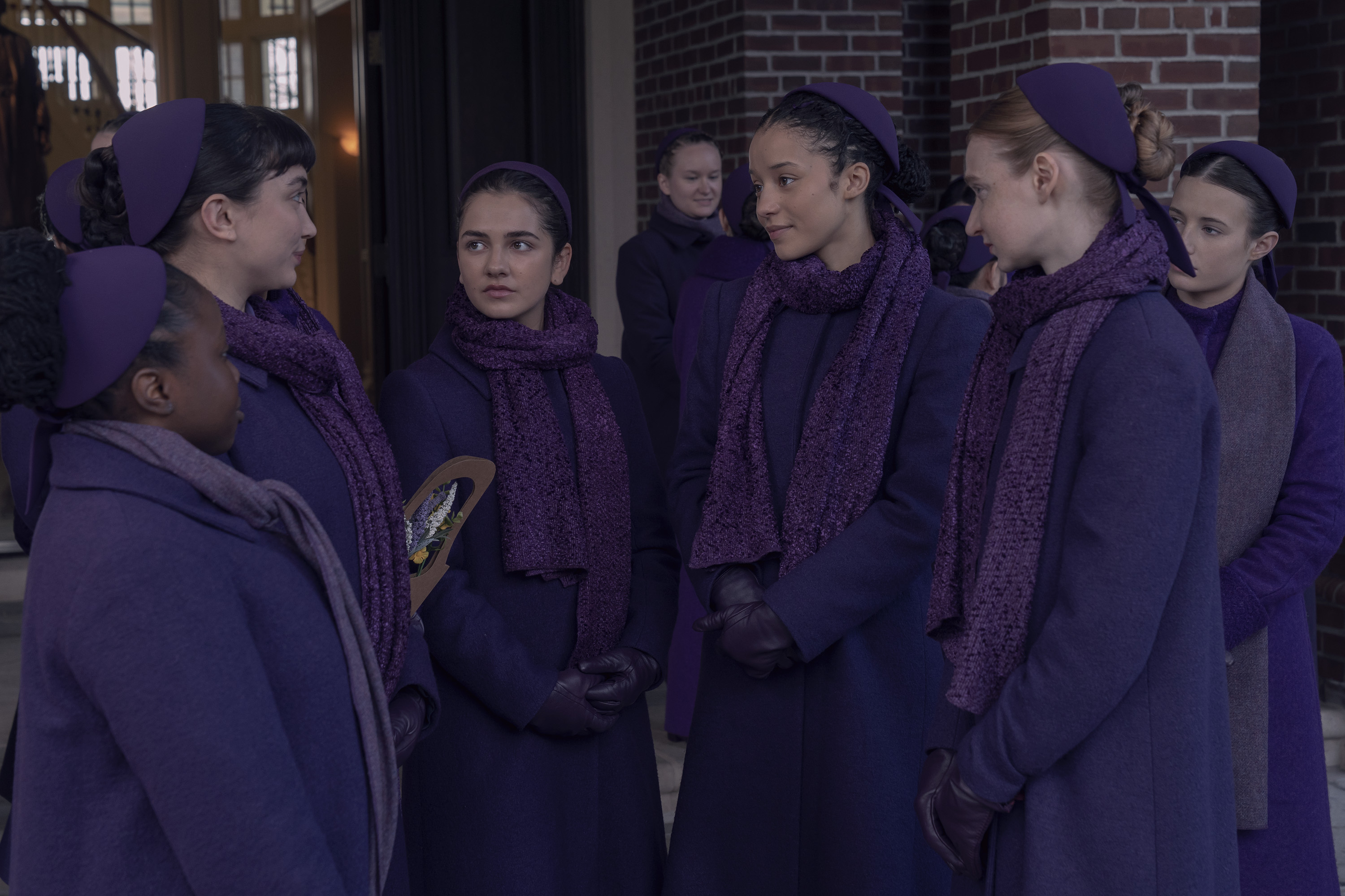 The ensemble cast of 'The Testaments' stand together dressed in purple outside of school