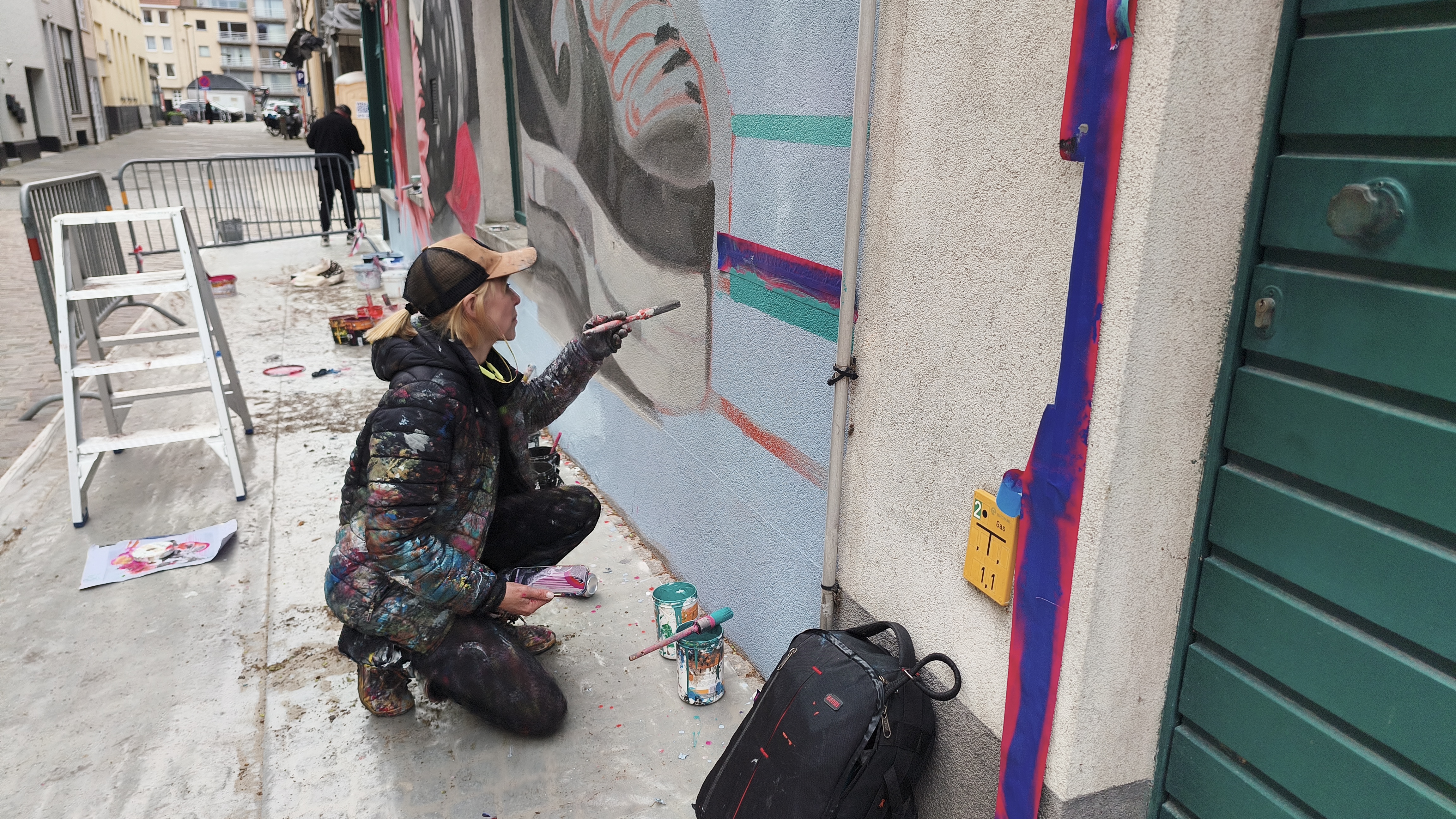 Artist Lula Goce in paint-splattered clothing crouches on a sidewalk while painting a detailed outdoor mural with a small brush.