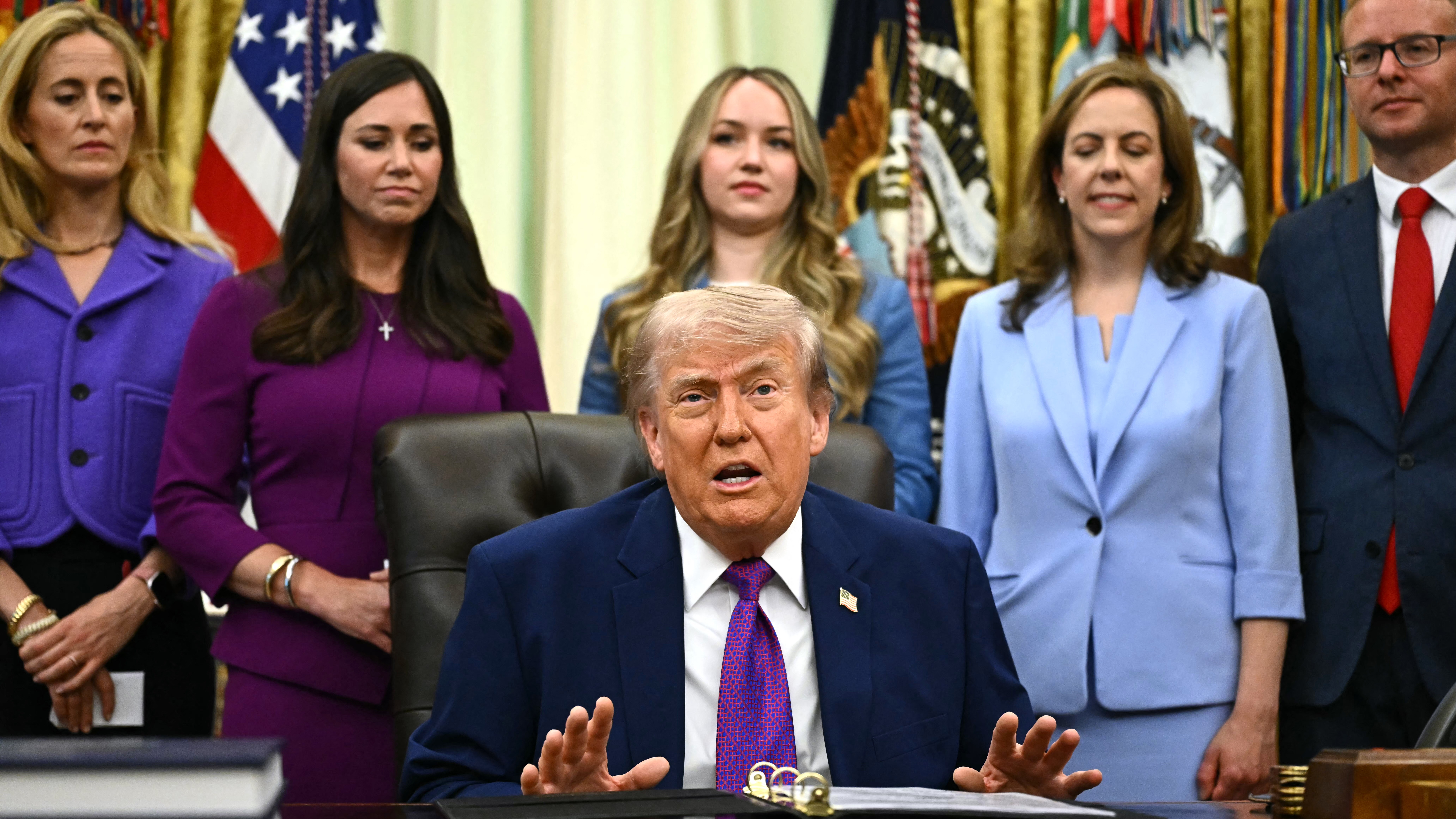 Trump is flanked by five people as he sits at his desk at the Oval Office.