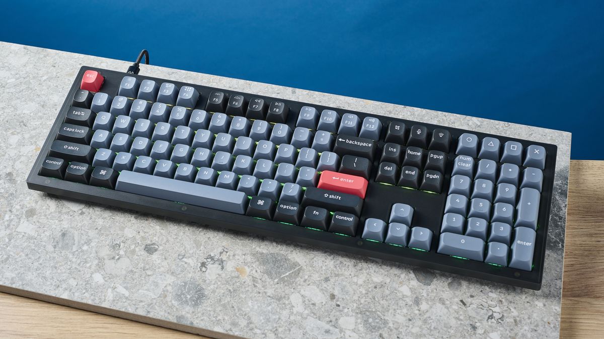 A photograph of the Keychron V6 in black, with gray , black and orange keycaps. The keyboard is positioned on a stone slate, with a blue wall in the background.