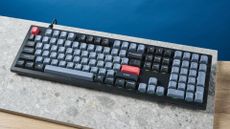 A photograph of the Keychron V6 in black, with gray , black and orange keycaps. The keyboard is positioned on a stone slate, with a blue wall in the background.