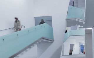 Blurred image of one person coming down stairs and another person at the top of the staircase. The staircase has glass banisters and the walls in the space is all white. There is a peek of the upper stair case above.