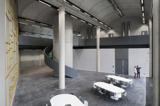 An open space in the building interior with high ceilings held up by white pillars, grey floors and grey spiral stairs that goes around a pillar. 3 white capsule shaped tables with matching chairs